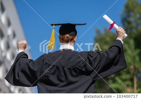 Rear view of a caucasian woman in a graduate gown holding a bundle with a diploma and standing near the university. Rear view of a caucasian woman in a graduate gown holding a bundle with a diploma and standing near the university. 103400717