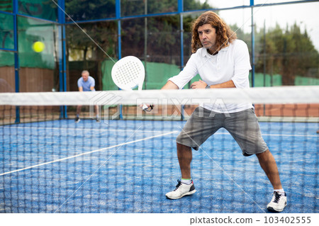 Portrait of middle-aged Latin male in sportswear enjoying popular sport padel game on tennis court outside 103402555