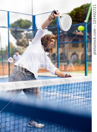 Focused middle-aged Latin man padel player hitting ball with racket on hard court in summer Focused middle-aged Latin man padel player hitting ball with racket on hard court in summer 103402721