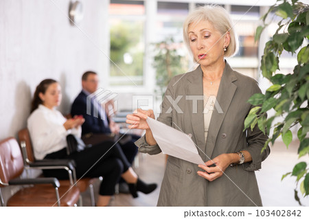Upset elderly woman with documents standing in reception 103402842