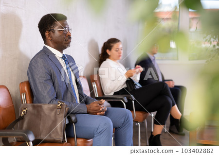 Serious-minded African male candidate siting on chair and waiting for job interview in hallway Serious-minded African male candidate siting on chair and waiting for job interview in hallway 103402882