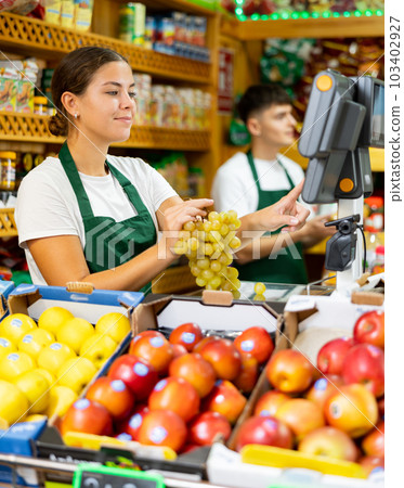 Young saleswoman weighs grapes on a scale Young saleswoman weighs grapes on a scale 103402927