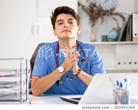 Portrait of man doctor in surgical scrubs sitting at working table in office and using laptop Portrait of man doctor in surgical scrubs sitting at working table in office and using laptop 103403169