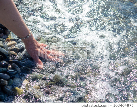 A hand with a bracelet made of stones reaches for the water. Sea wave and female hand. Feel the wave. Pebble beach. A hand with a bracelet made of stones reaches for the water. Sea wave and female hand. Feel the wave. Pebble beach. 103404975