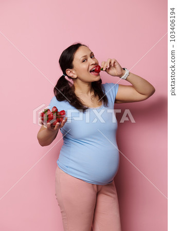 Gorgeous pregnant woman eating fresh organic strawberries, isolated over pink background. Expectant mother in blue t-shirt, holding bowl with fresh berries. Healthy nutrition and pregnancy concept. Gorgeous pregnant woman eating fresh organic strawberries, isolated over pink background. Expectant mother in blue t-shirt, holding bowl with fresh berries. Healthy nutrition and pregnancy concept. 103406194