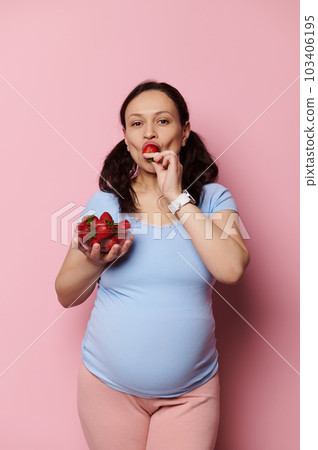 Vertical advertising studio shot on pink background of an expectant pregnant mother in blue t-shirt, looking at camera, holding a bowl with fresh berries and tasting yummy strawberries. Healthy diet Vertical advertising studio shot on pink background of an expectant pregnant mother in blue t-shirt, looking at camera, holding a bowl with fresh berries and tasting yummy strawberries. Healthy diet 103406195