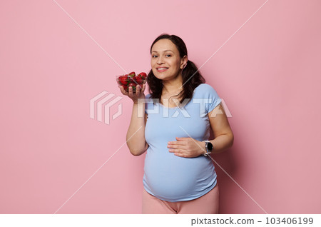 Happy young pregnant woman in blue casual t-shirt, holding bowl with fresh ripe organic strawberries, smiling looking at camera, isolated on pink. Healthy nutrition and balanced diet in pregnancy time 103406199