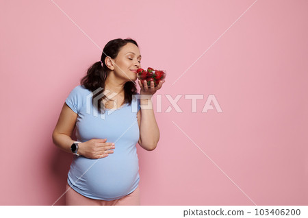 Attractive conscious pregnant woman puts her hand on her big belly in late pregnancy, enjoying the smell of delicious organic strawberries in a glass bowl, isolated on pink background. Copy ad space Attractive conscious pregnant woman puts her hand on her big belly in late pregnancy, enjoying the smell of delicious organic strawberries in a glass bowl, isolated on pink background. Copy ad space 103406200