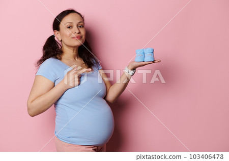Beautiful Latina pregnant woman in blue t-shirt, looking at camera, pointing finger aside cute baby booties on her hand, isolated on pink background. Happy mother expecting baby. Pregnancy. Maternity 103406478