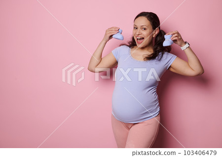 Happy pregnant woman holds blue knitted baby booties, smiles broadly looking at camera, isolated on pink background. Expectant mother shares positive emotions of her pregnancy and maternity lifestyle Happy pregnant woman holds blue knitted baby booties, smiles broadly looking at camera, isolated on pink background. Expectant mother shares positive emotions of her pregnancy and maternity lifestyle 103406479