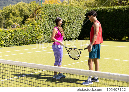 Side view of biracial young couple holding tennis rackets while standing in court against plants 103407122