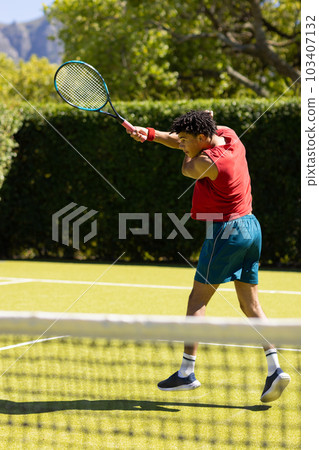 Biracial young man hitting ball with tennis racket while playing at tennis court on sunny day 103407132