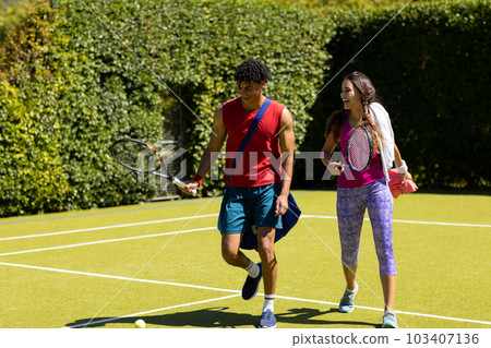Cheerful biracial young couple with rackets walking out of tennis court after playing game Cheerful biracial young couple with rackets walking out of tennis court after playing game 103407136