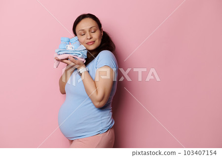 Studio portrait of happy gravid pregnant woman touching her face to the clothes of future newborn baby, posing with eyes closed on pink background. Copy space. Pregnancy fashion. Maternity. Childbirth Studio portrait of happy gravid pregnant woman touching her face to the clothes of future newborn baby, posing with eyes closed on pink background. Copy space. Pregnancy fashion. Maternity. Childbirth 103407154