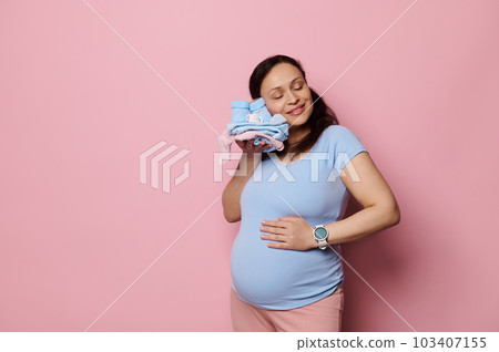 Happy pregnant woman, gently caressing her belly, touches her face to the clothes of future baby, enjoying her pregnancy and maternal lifestyle, expressing positive emotions, isolated pink background 103407155