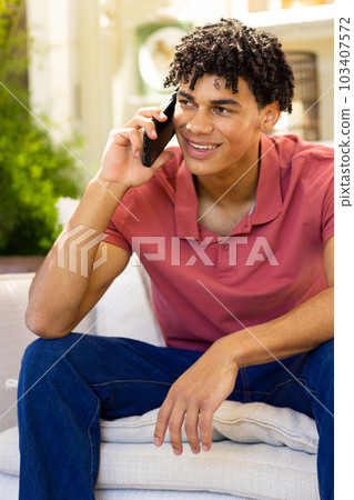 Biracial smiling young man talking over smartphone while sitting on sofa in living room 103407572