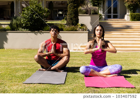 Biracial young couple practicing meditation in prayer pose while sitting on mats in yard Biracial young couple practicing meditation in prayer pose while sitting on mats in yard 103407592