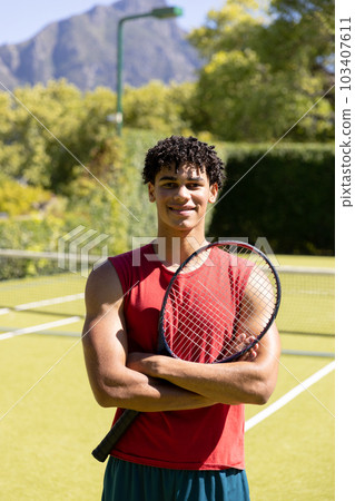 Smiling biracial young man with arms crossed holding tennis racket while standing in court 103407611