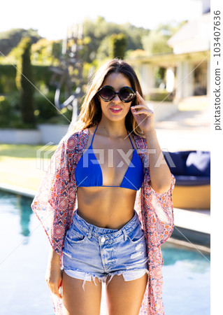 Portrait of pretty biracial young woman in sunglasses and bikini standing at poolside against sky Portrait of pretty biracial young woman in sunglasses and bikini standing at poolside against sky 103407636