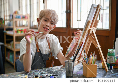 A happy Asian gay art student s painting watercolors on a small canvas at his desk A happy Asian gay art student s painting watercolors on a small canvas at his desk 103409341
