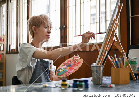 A creative Asian gay man or queer art student is painting watercolors on a small canvas A creative Asian gay man or queer art student is painting watercolors on a small canvas 103409342