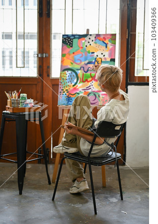 Back view. A talented young Asian queer artist sits on a chair and paints his picture on a canvaS Back view. A talented young Asian queer artist sits on a chair and paints his picture on a canvaS 103409366