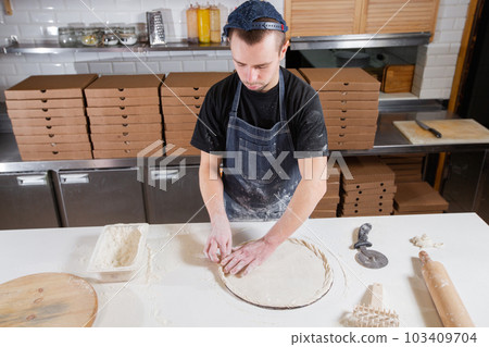 The chef prepares pizza. Raw pizza ready to bake. Cook in a blue apron in the kitchen. with a shovel in his hands. boxes for food delivery on background. 103409704