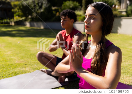 Biracial young woman with boyfriend practicing meditation in prayer pose while sitting in yard 103409705