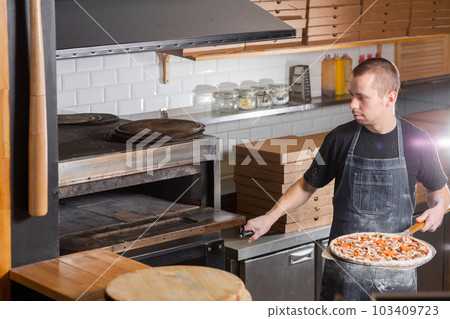 The chef prepares pizza. Raw pizza ready to bake. Cook in a blue apron in the kitchen. with a shovel in his hands. boxes for food delivery on background. 103409723