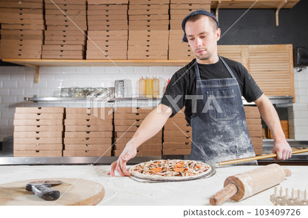 The chef prepares pizza. Raw pizza ready to bake. Cook in a blue apron in the kitchen. with a shovel in his hands. boxes for food delivery on background. The chef prepares pizza. Raw pizza ready to bake. Cook in a blue apron in the kitchen. with a shovel in his hands. boxes for food delivery on background. 103409726