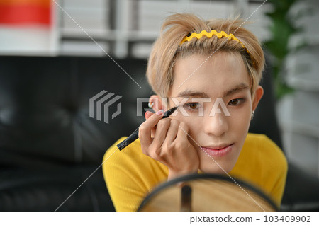 A gay man applies liquid eyeliner to create a winged look while doing his makeup at home A gay man applies liquid eyeliner to create a winged look while doing his makeup at home 103409902