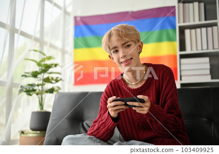 A smiling young Asian gay man sits on a sofa with his smartphone in his hands A smiling young Asian gay man sits on a sofa with his smartphone in his hands 103409925