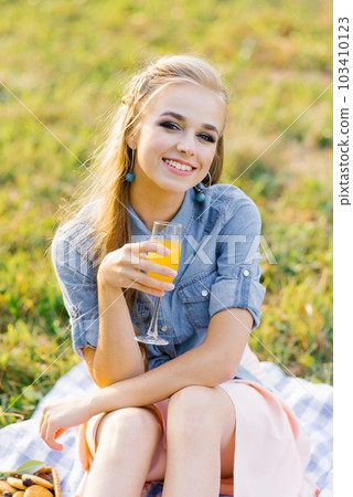 Young woman in a blue denim shirt and pink skirt in the garden at a picnic holding a glass of juice in her hand Young woman in a blue denim shirt and pink skirt in the garden at a picnic holding a glass of juice in her hand 103410123