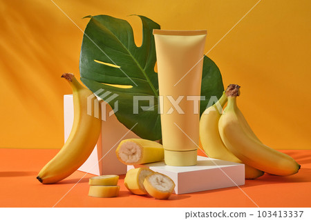 A yellow cosmetic tube on white podium, decorated with fresh bananas and jungle monstera leaves on a yellow background with leaf shadow. Mockup scene for advertising cosmetic of banana extract A yellow cosmetic tube on white podium, decorated with fresh bananas and jungle monstera leaves on a yellow background with leaf shadow. Mockup scene for advertising cosmetic of banana extract 103413337
