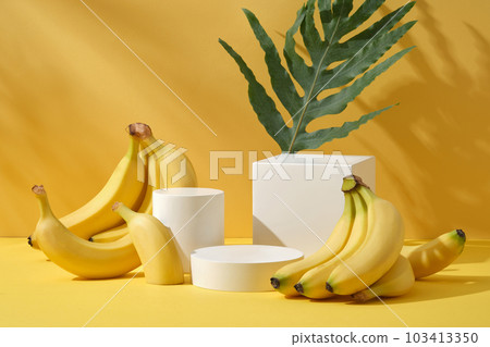 Bunch of bright yellow tiny bananas decorated on a yellow background with cylinder white podiums and green leaf. Minimal scene for cosmetic product presentation. Front view 103413350