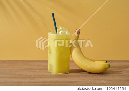 Front view of glass of banana juice and ripe yellow banana placed on wooden table on yellow background with palm leaf shadow. Scene for advertising product from banana with copy space Front view of glass of banana juice and ripe yellow banana placed on wooden table on yellow background with palm leaf shadow. Scene for advertising product from banana with copy space 103413364