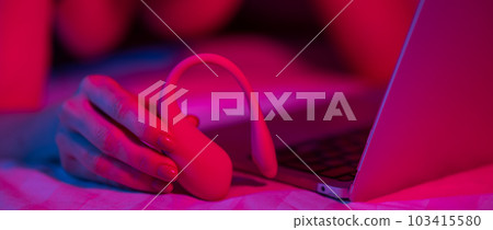 A woman is lying in bed holding a curved vibrator and looking at a laptop. Girl using sex toy in blue-red light. A woman is lying in bed holding a curved vibrator and looking at a laptop. Girl using sex toy in blue-red light. 103415580