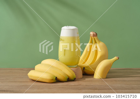 A glass of banana juice placed on a wooden podium, decorated with bunches of banana. Bananas contains an abundance of vitamin A, which restores dull skin and helps brighten skin. 103415710