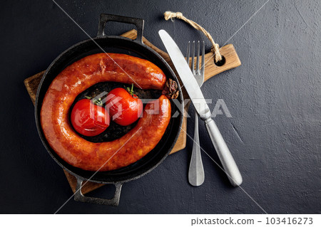 Grilled pork sausage in a cast iron skillet with tomatoes and cutlery on a wooden cutting board on a dark stone table 103416273
