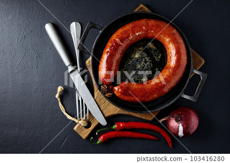 Grilled sausage rolled into a ring on a cast-iron pan with cutlery, a wooden cutting board, hot peppers and red onions on a black stone background Grilled sausage rolled into a ring on a cast-iron pan with cutlery, a wooden cutting board, hot peppers and red onions on a black stone background 103416280