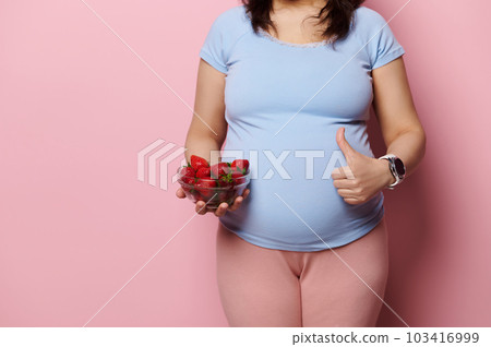 Cropped view of the belly of pregnant woman gesturing with thumb up, holding a glass bowl with fresh ripe strawberries, isolated on pink background. The concept of healthy nutrition in pregnancy time Cropped view of the belly of pregnant woman gesturing with thumb up, holding a glass bowl with fresh ripe strawberries, isolated on pink background. The concept of healthy nutrition in pregnancy time 103416999