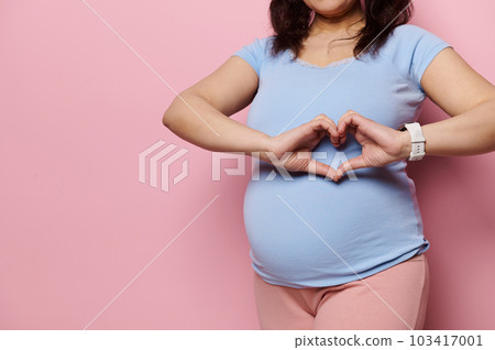 Focus on the hands of a pregnant woman, expectant gravid mother in blue t-shirt, showing heart shape made with her fingers over her big belly, expressing love, tenderness, isolated on pink background 103417001