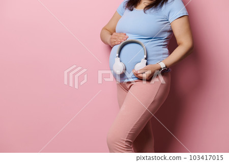 Close-up studio shot of pregnant woman putting wireless headphones over her belly, turning on soothing music, feeling connection with her baby in womb, isolated on pink background. Pregnancy lifestyle 103417015