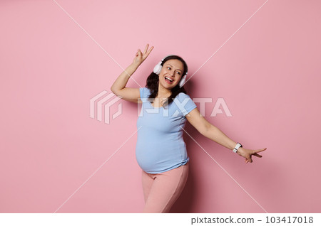 Joyful Latin American pregnant woman listening to music in wireless headphones, smiling looking aside, dancing over pink studio background. Copy ad space. Expectant mother enjoying maternity lifestyle 103417018