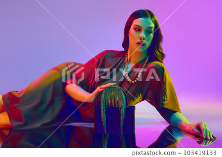 Portrait with attractive fashion model wearing trendy clothes with a silk dress and accessories over violet background in neon light 103419133
