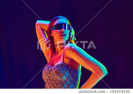 High fashion model wearing futuristic style of clothes with black sunglasses posing over violet studio background in neon light. Close up High fashion model wearing futuristic style of clothes with black sunglasses posing over violet studio background in neon light. Close up 103419140