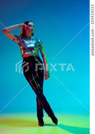 High fashion model, young woman wearing trendy clothes with leather black pants and silver top posing with over blue background in neon light. Concept of beauty, fashion week High fashion model, young woman wearing trendy clothes with leather black pants and silver top posing with over blue background in neon light. Concept of beauty, fashion week 103419153
