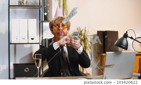Cheerful rich business man working on office laptop wasting throwing money to camera profit win 103420622