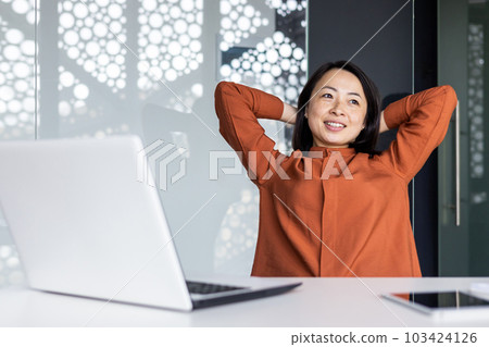 Happy and smiling successful Asian business woman finished work satisfied with work result and achievement, hands behind head and smiling close up 103424126