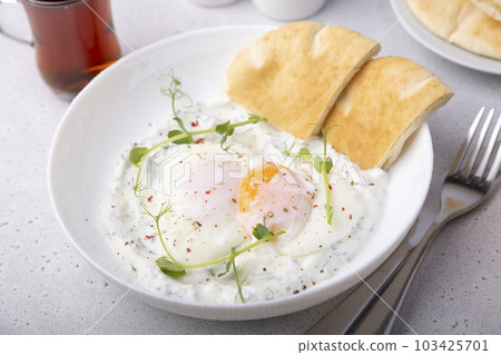 Turkish eggs (chilbir) with yogurt, fragrant butter, pea sprouts, flatbread and tea. Traditional Turkish breakfast with poached eggs (with liquid yolk). Selective focus, close-up. 103425701